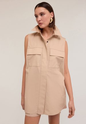 Abito chemisier senza maniche color beige realizzato in tessuto morbido, con colletto alto, due tasche frontali e una chiusura frontale con bottoni.