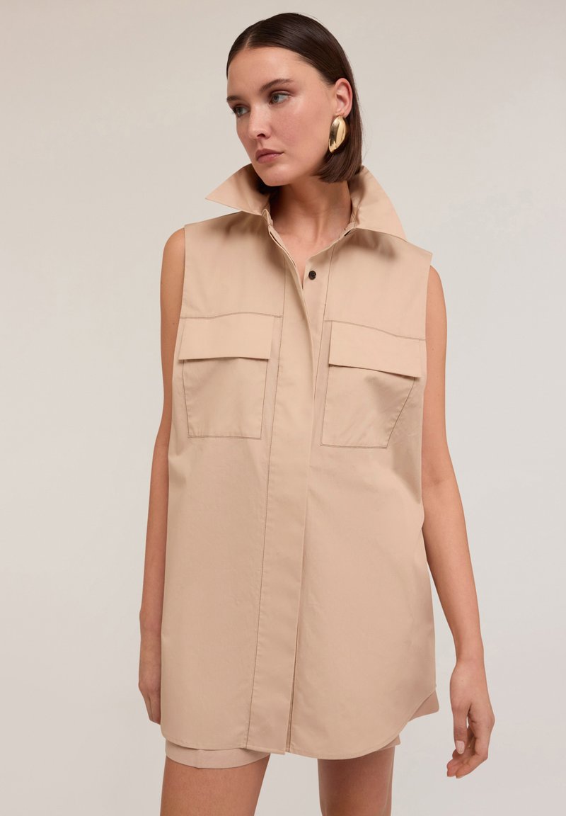 Vestido sin mangas de color beige hecho de una tela suave, con un alto cuello, dos bolsillos frontales y una botonadura en la parte delantera.