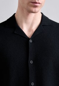 Melnā, teksturēta džemperis ar mīkstu apdari, piedāvā kolu piegriezumu un četras melnas pogas priekšpusē.