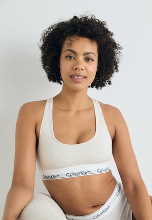 Lys grå bomulls-bralette med bred hvit stropp som har "Calvin Klein"-tekst. Myk tekstur, firkantet utringning og racerback-design.
