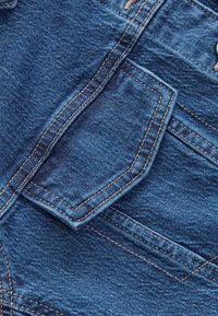 Közeli kép kék farmer anyagról, részletes varrással és hajtogatott háromszög alakú zsebfedéllel.