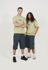 Duos modeliai vilki derančius šviesiai žalius grafinius marškinėlius su juodais ir mėlynais raštais, derinamus su tamsiai mėlynomis džinsinėmis šortais ir juodai baltais sportiniais bateliais.