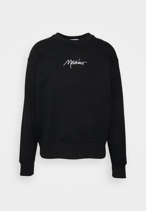 Henkilö, jolla on päällä musta Moschino-teksti-containing collegepaita, mustat nahkahousut ja mustavalkoiset nauhalliset kengät, seisoo yksiväristä taustaa vasten.