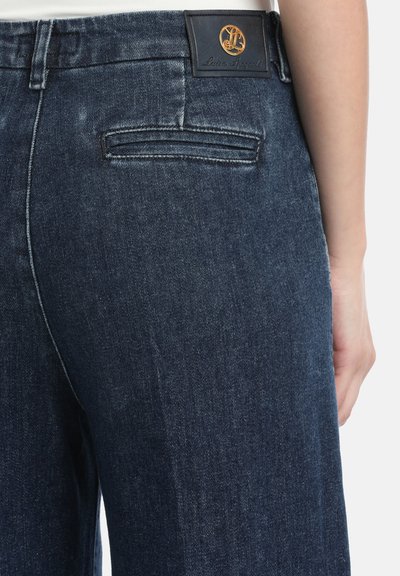 Close-up van donkerblauwe denim broek gedragen door een persoon, met zicht op de details van het achterzak en een zwart patch met gouden en leren branding.