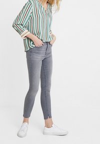 Blusa a rayas en verde, blanco y marrón, con mangas largas y escote en pico. Combinada con jeans gris ajustados y zapatillas blancas.