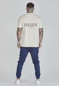 Kermanen puuvillainen t-paita lyhyillä hihoilla, jossa on ruskea "SIKSILK" -logo ja teksti selässä, yhdistettynä tummansinisiin kapealahkeisiin farkkuihin ja valkoisiin kenkiin.