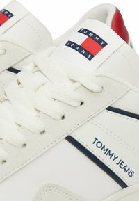Fehér sportcipők textil felsőrésszel, texturált részletekkel, kék és piros márkázott részlettel, lapos fehér fűzőkkel és párnázott bokarésszel.