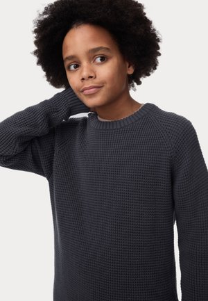 Barn med krøllet hår iført en mørkegrå tekstureret striktrøje, der løfter en arm bag hovedet og ser let til siden.