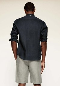 Chemise bleu foncé à manches longues avec poignets retroussés et détail de bouton au col, associée à un short gris clair. Présente une texture de tissu lisse.