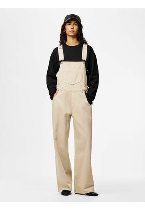 Oseba stoji, oblečena v bež široke hlače na naramnice, črno majico z dolgimi rokavi, črno kapo in črne čevlje, roke v žepih, nevtralen ozadje.