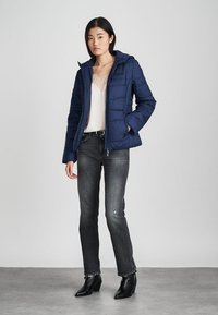 Mörkblå pufferjacka med huva, har quiltad textur, dragkedja och sidofickor, bärs över en krämfärgad topp och mörka jeans.