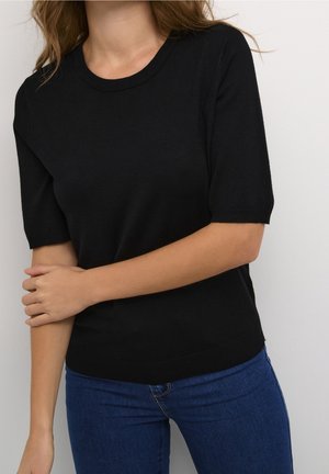 Femme portant un top en maille noir ajusté à manches courtes et un jean bleu, un bras croisé sur l'autre devant un fond uni.