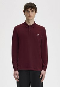 Polo a maniche lunghe in cotone bordeaux, con colletto, pattina con tre bottoni e un piccolo emblema bianco sul lato sinistro del petto.