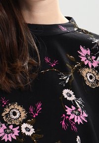 Tissu noir avec un motif floral comportant des fleurs roses, blanches et beiges. Encolure ronde avec une texture subtile. Design près du corps.