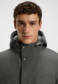 Grau Jacke mit hohem Kragen, die eine Kapuze, Druckknöpfe und eine glatte Textur aufweist. Enthält metallische Akzente und ein minimalistisches Design.