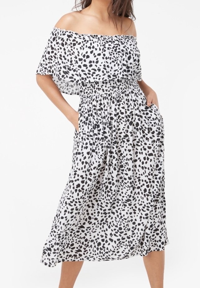 Femme portant une robe midi blanche à épaules dénudées avec des taches noires irrégulières et les mains dans les poches latérales.