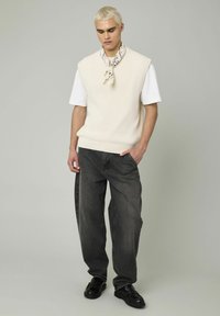 Un pull sans manches crème porté sur un t-shirt blanc, associé à un jean baggy gris foncé et des mocassins noirs. Comprend une écharpe à motifs.