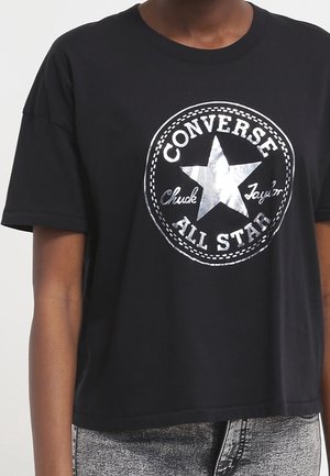 Personne portant un t-shirt court noir avec un logo Converse All Star argenté et un jean gris délavé acide.