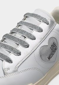 Άσπρο αθλητικό παπούτσι με ασημί κορδόνια με υφή και μεταλλική πλάκα, που διαθέτει έμπλαστρο σε σχήμα καρδιάς με το κείμενο «Love Moschino».