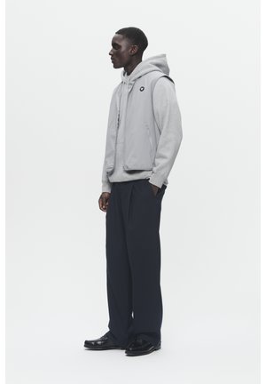 Gray sleeveless vest over gray hoodie, paired with loose black pants and shiny black shoes. Vest features side pockets and logo accent.