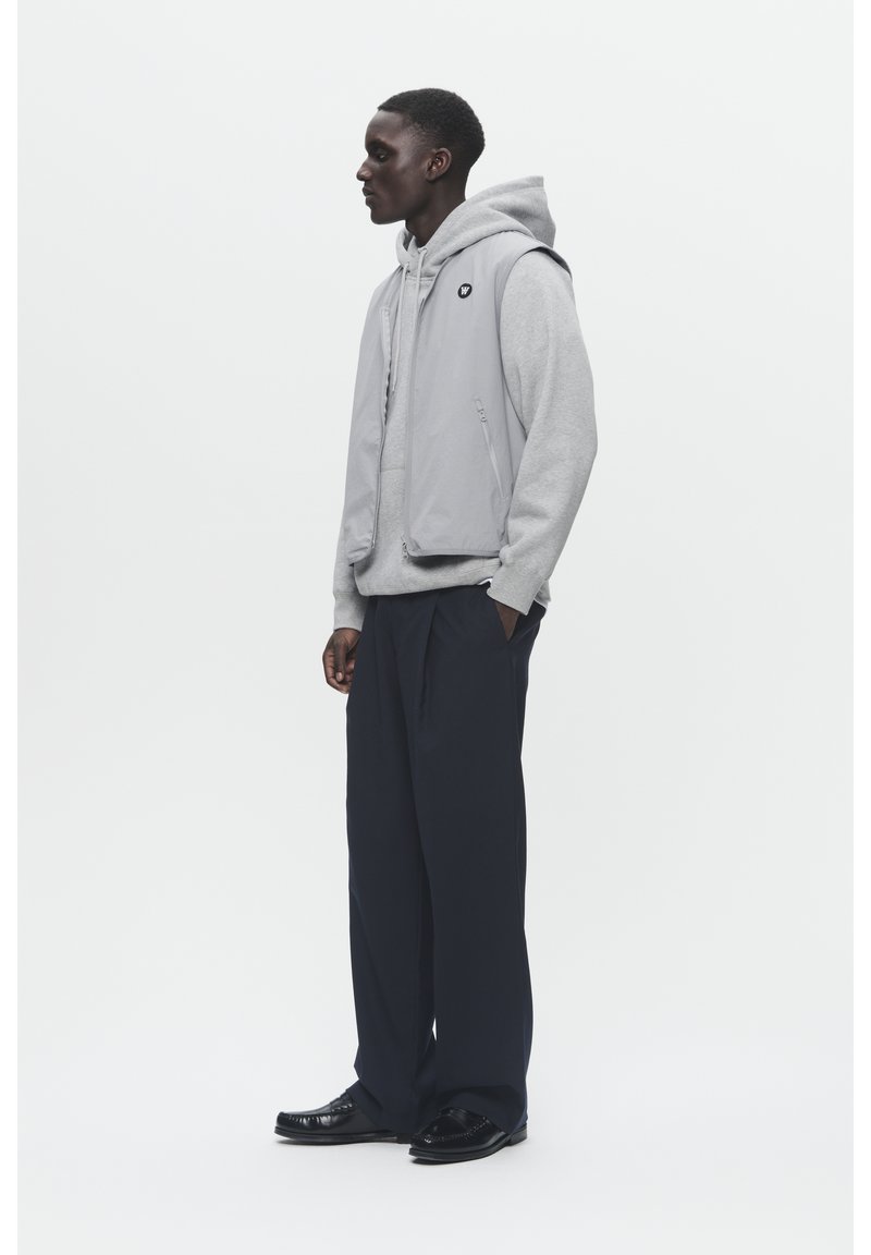 Grijze mouwloze vest over een grijze hoodie, gecombineerd met losse zwarte broek en glanzende zwarte schoenen. Het vest heeft zijzakken en een logo-accent.