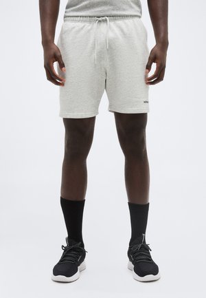 Bărbat purtând pantaloni scurți gri deschis cu șnur, șosete negre până la gleznă și încălțăminte sport neagră, stând în picioare pe un fundal simplu, deschis la culoare.