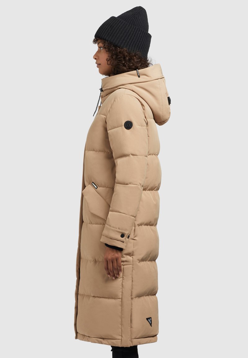 Doudoune beige avec capuche, design matelassé, poches latérales, patch noir sur la manche et silhouette épurée. Portée avec un bonnet noir.