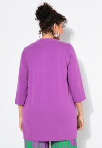 Lila Jersey-Tunika mit Dreiviertelärmel. Glatte Textur, lockerer Schnitt und abgerundeter Saum. Keine sichtbaren Muster oder Verzierungen.