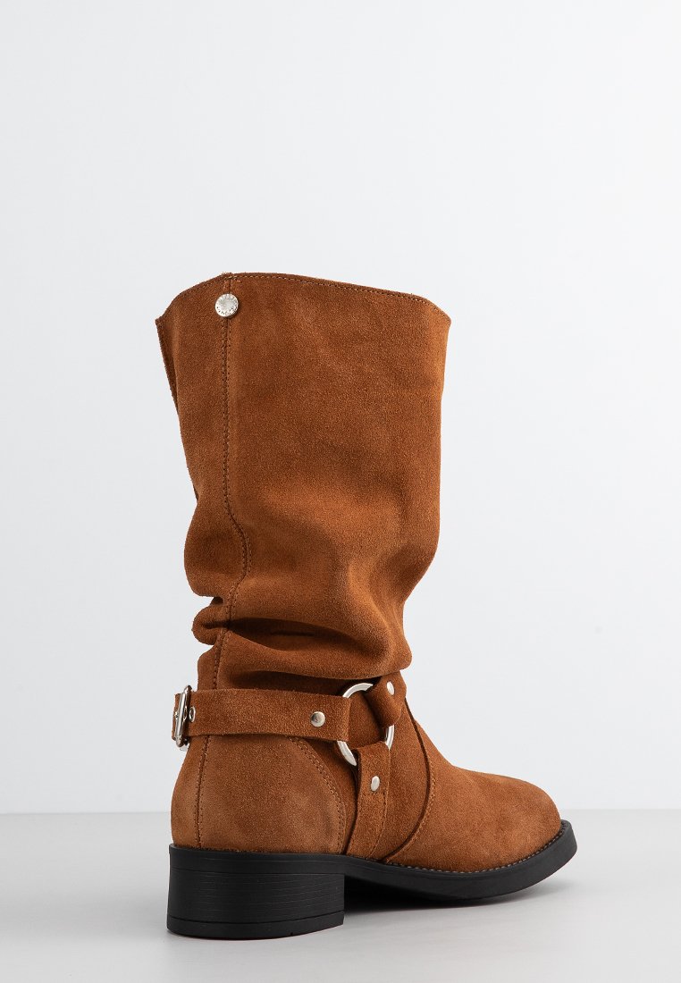 Braune Wildleder-Stiefel bis zur Wade mit schlaffer Schaft, silberner Schnalle und Ringdetails sowie niedrigem schwarzen Absatz auf einem schlichten weißen Hintergrund.