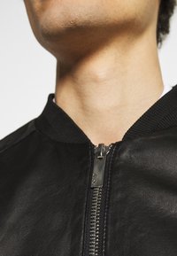 Svart läderbomberjacka med en stickad krage, prydd med en silverfärgad dragkedja och slät yta. Nära bild av detalj vid halslinningen.