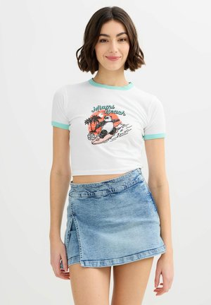 Joven con cabello corto y marrón que lleva un top corto blanco con un panda surfista y el texto "Miami Beach", combinado con una falda vaquera azul clara.