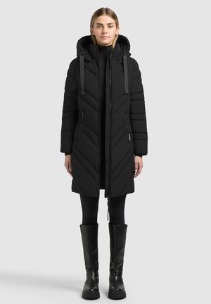 Femme portant un long manteau noir matelassé avec capuche, des leggings noirs et des bottes noires montantes jusqu'aux genoux, debout devant un fond clair uni.