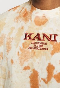 Triko s batikou v béžové a oranžové barvě s červeným vyšívaným textem "KANI THE ORIGINAL EST. 1989 REG. TRADEMARK" na hrudi poblíž výstřihu.