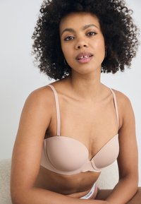 Beige underwire bra with smooth, molded cups, adjustable straps, and a soft elastic band featuring a subtle logo. Matte texture throughout.