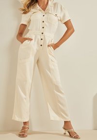 Krémový overal s krátkými rukávy, knoflíkovým zapínáním vpředu a dvěma velkými předními kapsami. Rozšířené nohavice a hladký textilní povrch. Minimalistický design.