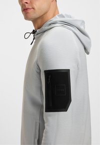 Férfi világosszürke kapucnis pulóverben, amely texturált anyagból készült, és fekete cipzáras folttal rendelkezik a felső ujján, rajta "BOSS" logóval.