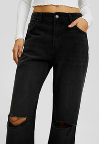 Musta denimi farkku, korkea vyötärö, jossa on kuluneet polvireiät, tavalliset taskut ja napitus. Pintakuvioitu, jossa on hieman kiiltoa.