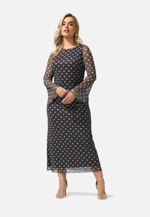 Femme portant une longue robe sombre à pois bleu clair et manches longues transparentes, debout, les mains jointes, portant des talons argentés.