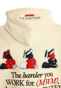 Sudadera con parches de zapatillas rojas y negras y el texto "Cuanto más duro TRABAJES por (MWM), mejor" bordado en tela beige.