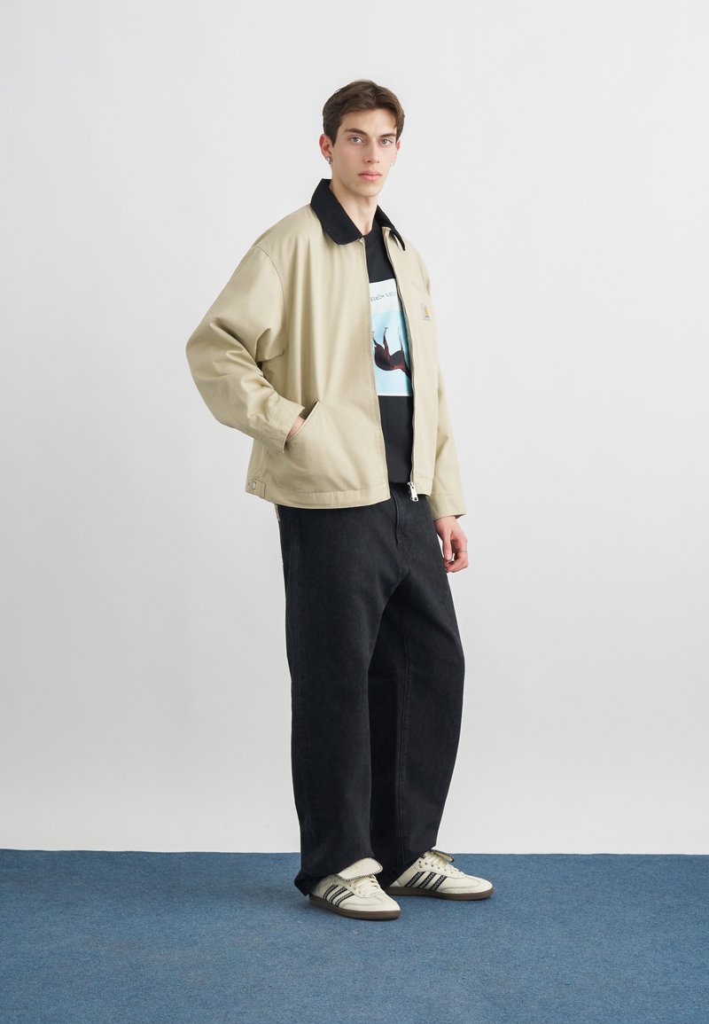 Joven de pie con una chaqueta beige con cuello negro, camiseta gráfica, pantalones negros holgados y zapatillas blancas con rayas negras.