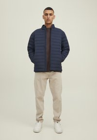Giacca imbottita blu navy con colletto alto e chiusura con zip, abbinata a pantaloni beige e sneakers bianche. Texture liscia e design trapuntato.