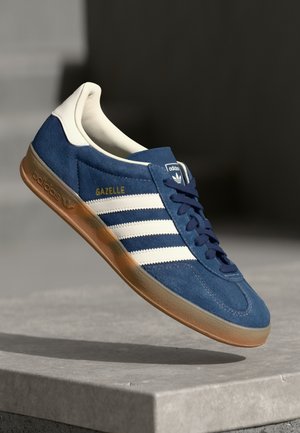 Blå Adidas Gazelle sneaker med hvide striber, beige inderside, gummisål og marineblå snørebånd, der svæver over en betongulv.