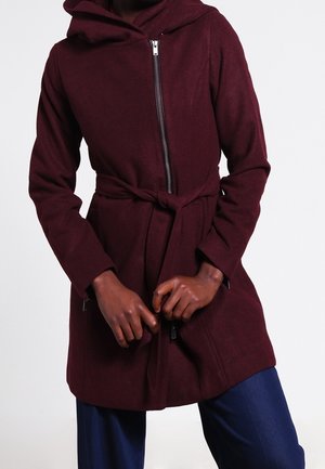 Man draagt een kastanjebruine jas met lange mouwen, rits en riem, gecombineerd met een blauwe broek, handen voor zich geklemd.