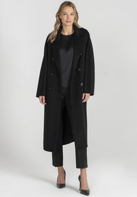 Czarny płaszcz wełniany z mieszanki włókien o podwójnym zapięciu, dużymi klapami i kieszeniami z przodu, w połączeniu z czarnymi spodniami i szpilkami.