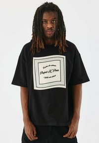 La camiseta negra de gran tamaño presenta un gráfico central en blanco con texto y un borde decorativo. Está hecha de suave tela de algodón y tiene mangas cortas.