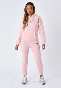 Felpa leggera rosa con tasca frontale e testo nero; pantaloni sportivi coordinati rosa chiaro con logo. Caratteristiche includono polsini elasticizzati e texture morbida.