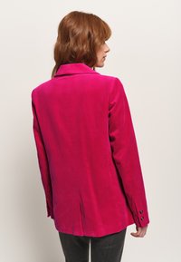Blazer in corduroy rosa brillante con colletto a rever, texture visibile, maniche lunghe e dettaglio sul retro a spacco, dotato di polsini con bottone.