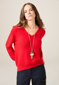 Czerwony sweter w serek wykonany z miękkiego materiału, po�łączony z ciemnymi dżinsami z misternymi ozdobami. Złoty naszyjnik z kolorowymi kamieniami.