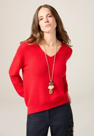 Czerwony sweter w serek wykonany z miękkiego materiału, połączony z ciemnymi dżinsami z misternymi ozdobami. Złoty naszyjnik z kolorowymi kamieniami.