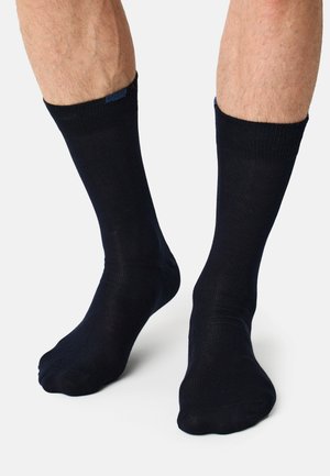 Ben iført ensfarvede sorte sokker til midt på læggen, stående på en hvid overflade, med synlig lys hud og benhår.